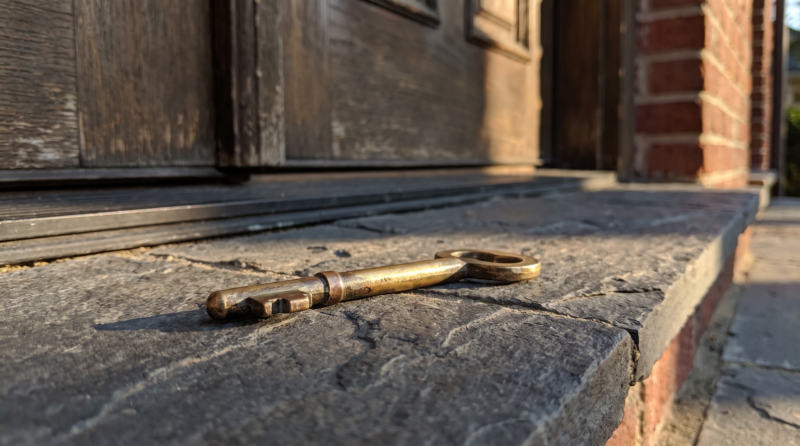 A brass door key resting on a stone entry threshold in warm afternoon light, with a brick home facade softly blurred behind it
