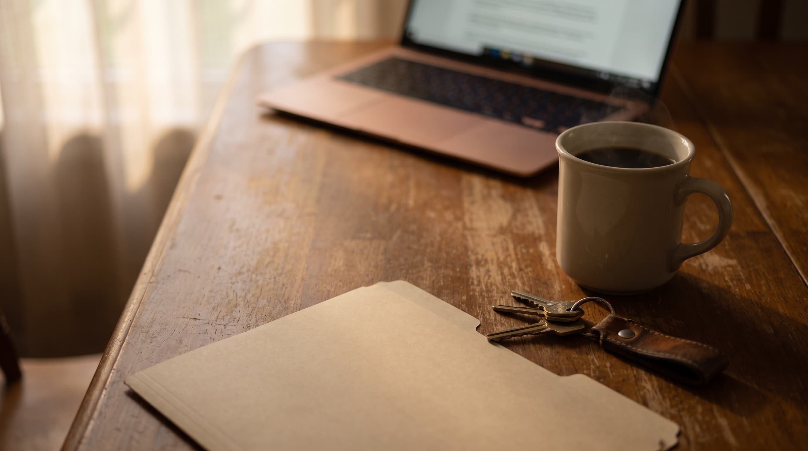 A closing folder and a set of house keys resting on a sunlit wood kitchen table beside a coffee mug, with a laptop glowing softly out of focus behind them.